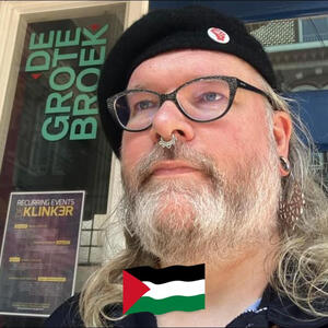 Selfie in front of De Klinker Selfie in front of De Klinker. I'm a white person with long grey hair and a grey beard. I'm wearing black rimmed glasses, a black beret with a little raised red fist pin on it, a septum piercing and large dangly earrings. Superimposed at the bottom is a sm
